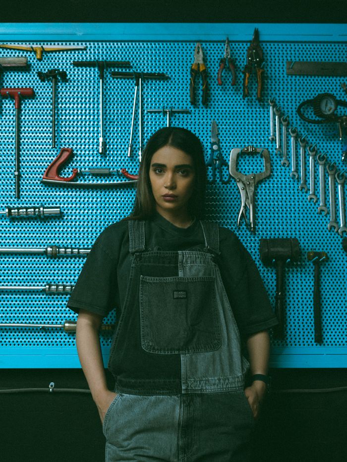 Young female mechanic in a workshop, standing against a tool wall wearing denim overalls.